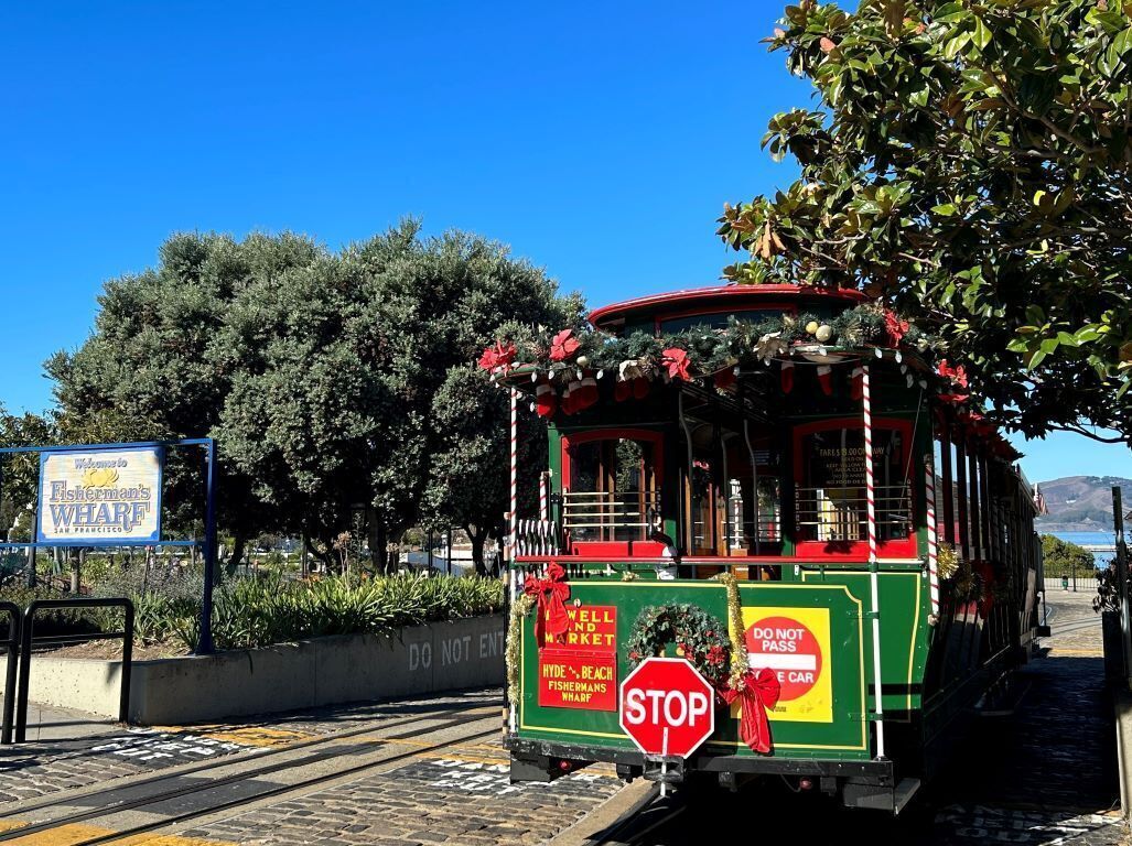 A green trolley with festive decorations, a stop sign, and a sign reading "Welcome to Fisherman's Wharf San Francisco".