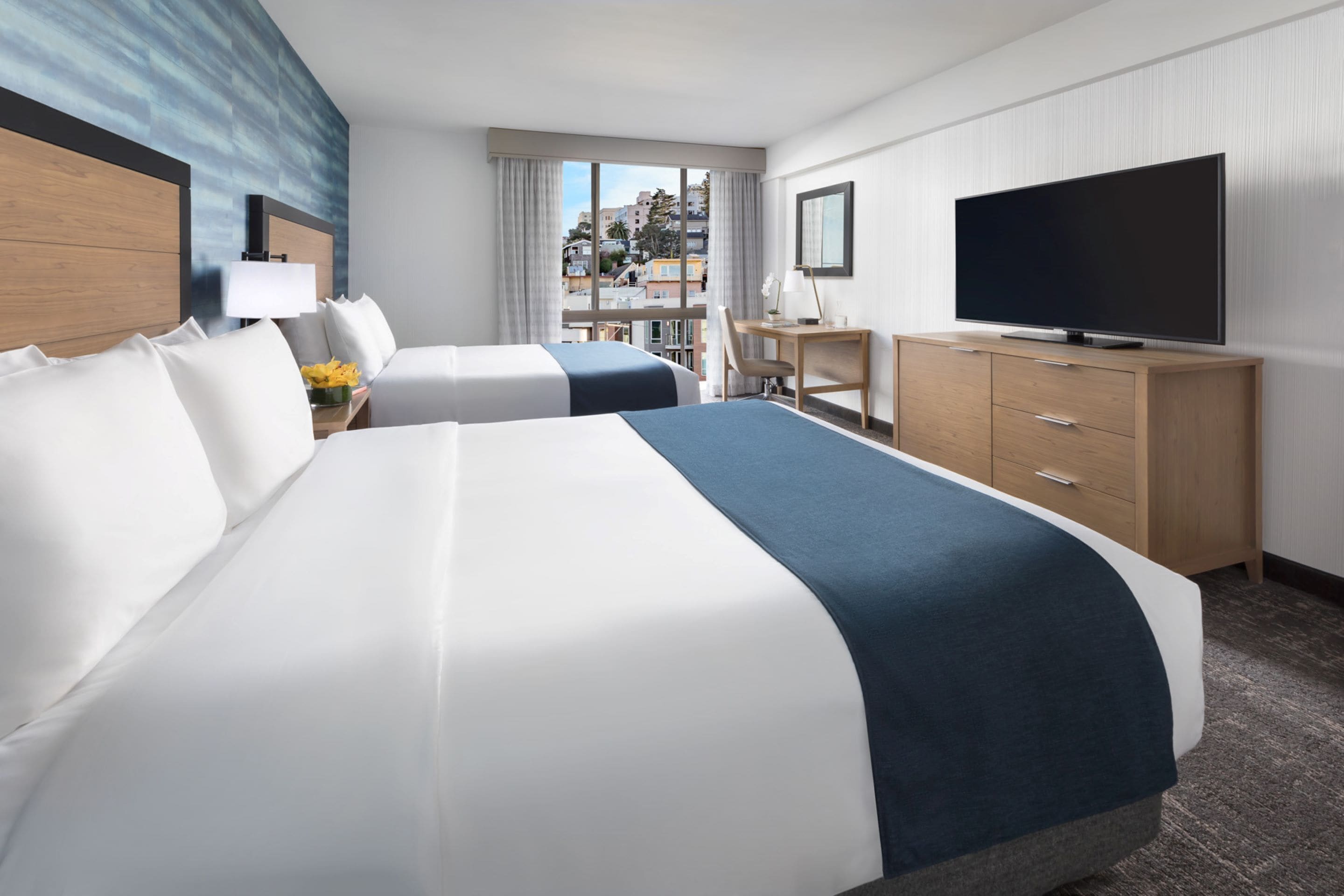 A hotel room with two beds, a desk, a TV, and a view of the city.