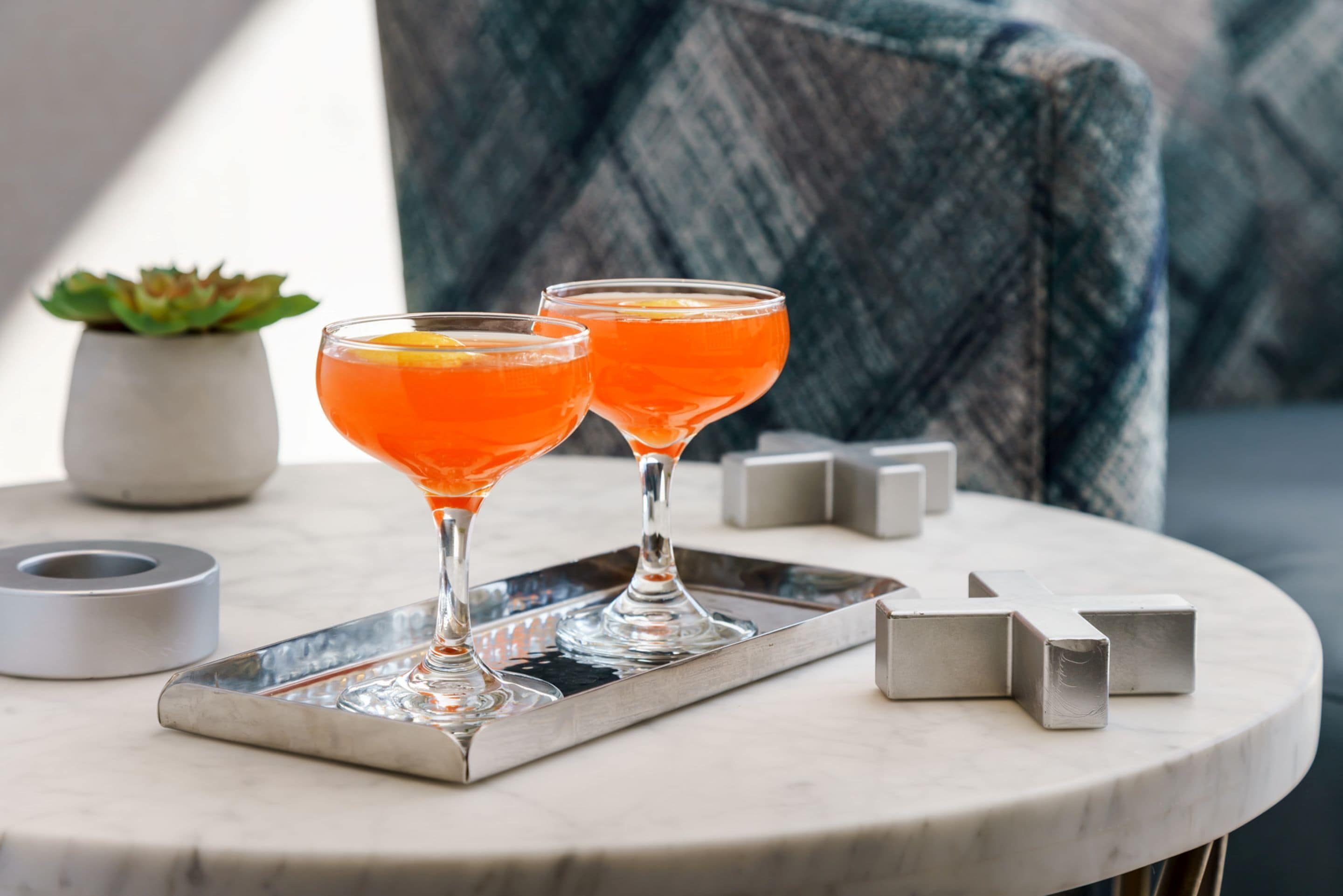To høje martini-glas med orange cocktails på et fad, en lille potteplante og geometriske metalblokke på et rundt bord.