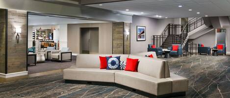A modern hotel lobby with a curved sofa, a staircase, and a dining area in the background.