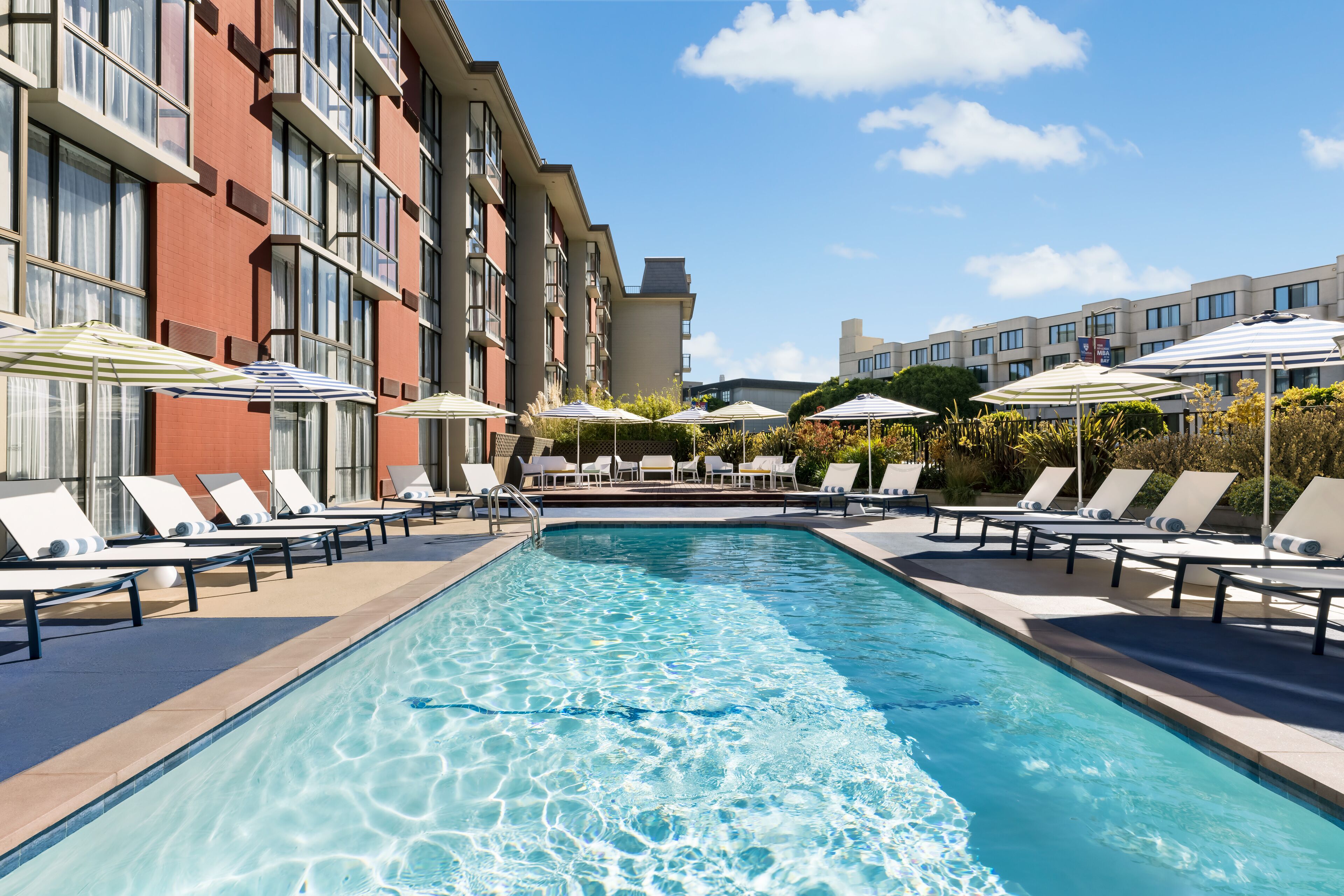 A swimming pool with lounge chairs and umbrellas, surrounded by a building with multiple floors and large windows.