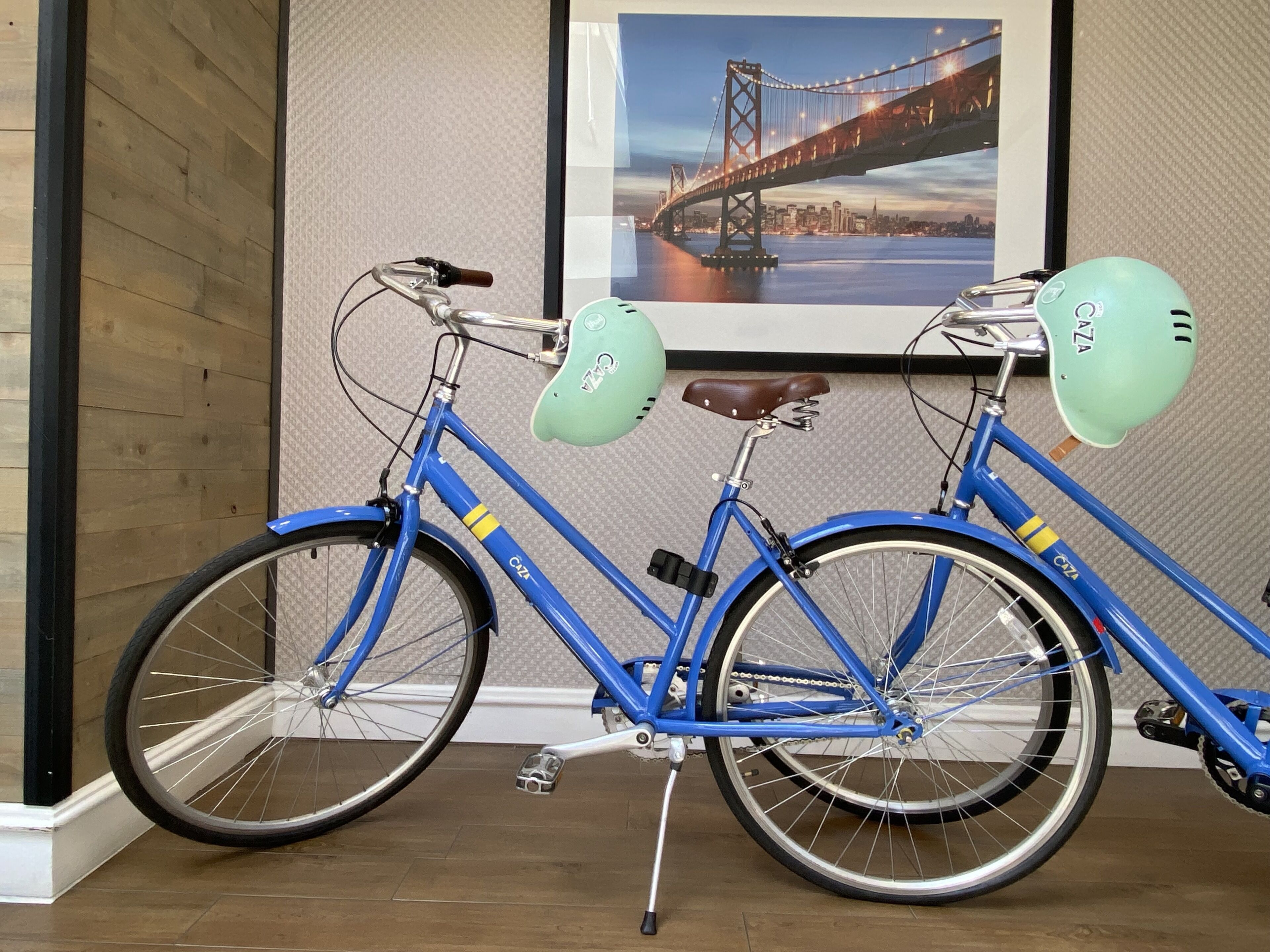 Una bicicleta azul con asiento marrón y dos cascos, apoyada contra una pared con un cuadro de un puente.