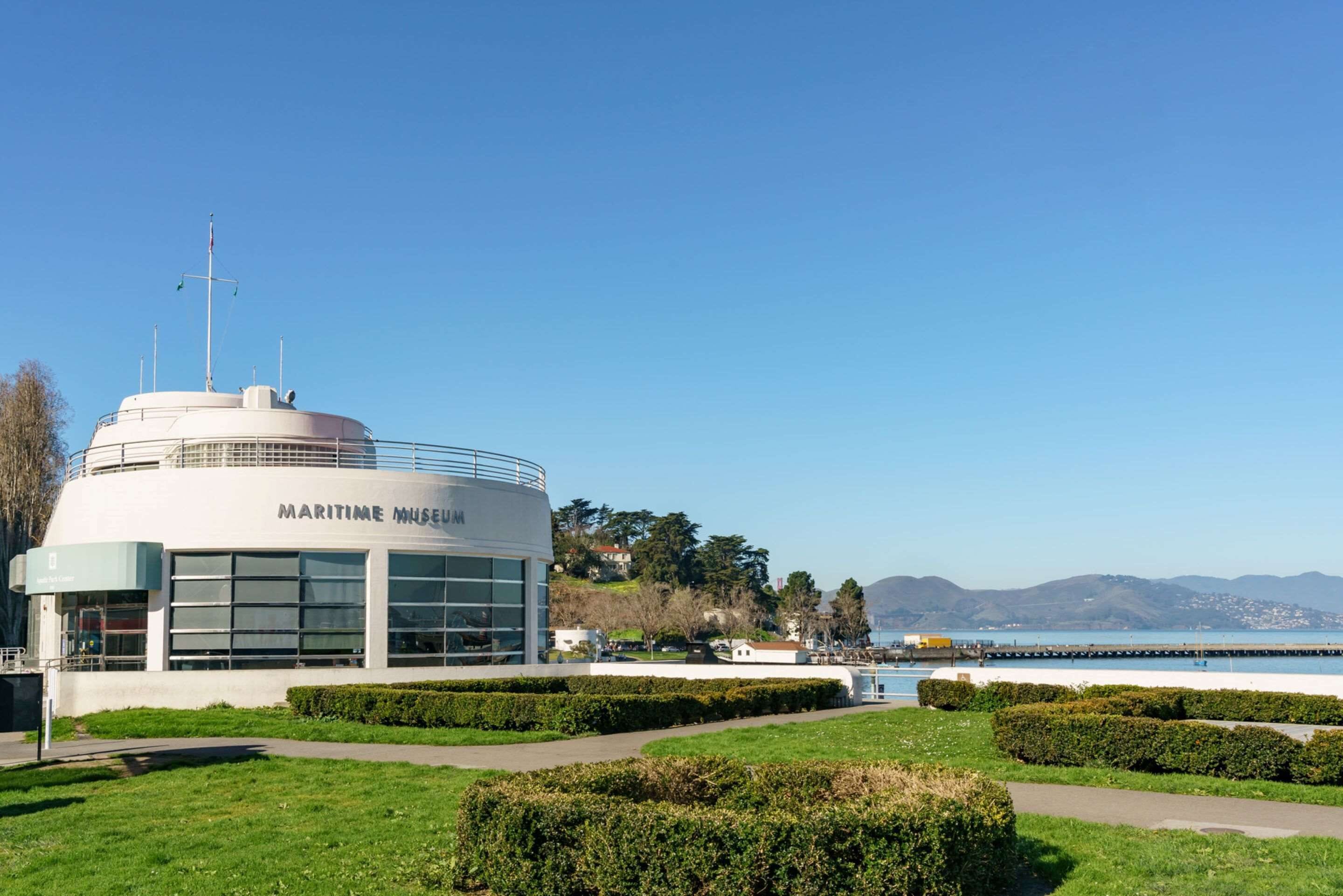 A maritime museum with a modern design, located near a waterfront with a clear view of the sea and hills in the background.
