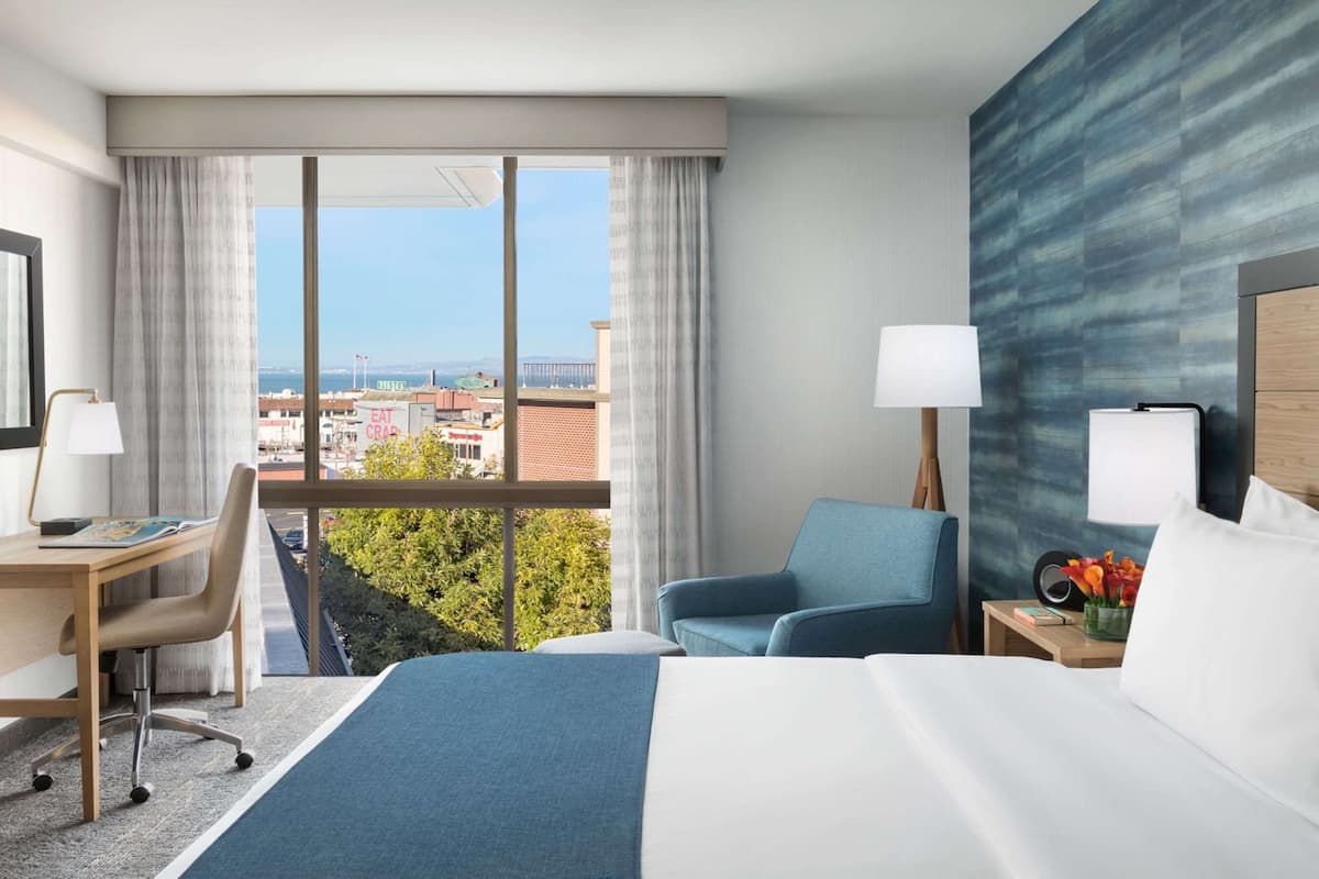 Una camera d'albergo con un letto grande, una scrivania con una sedia, una poltrona blu e vista sulla città attraverso ampie finestre.