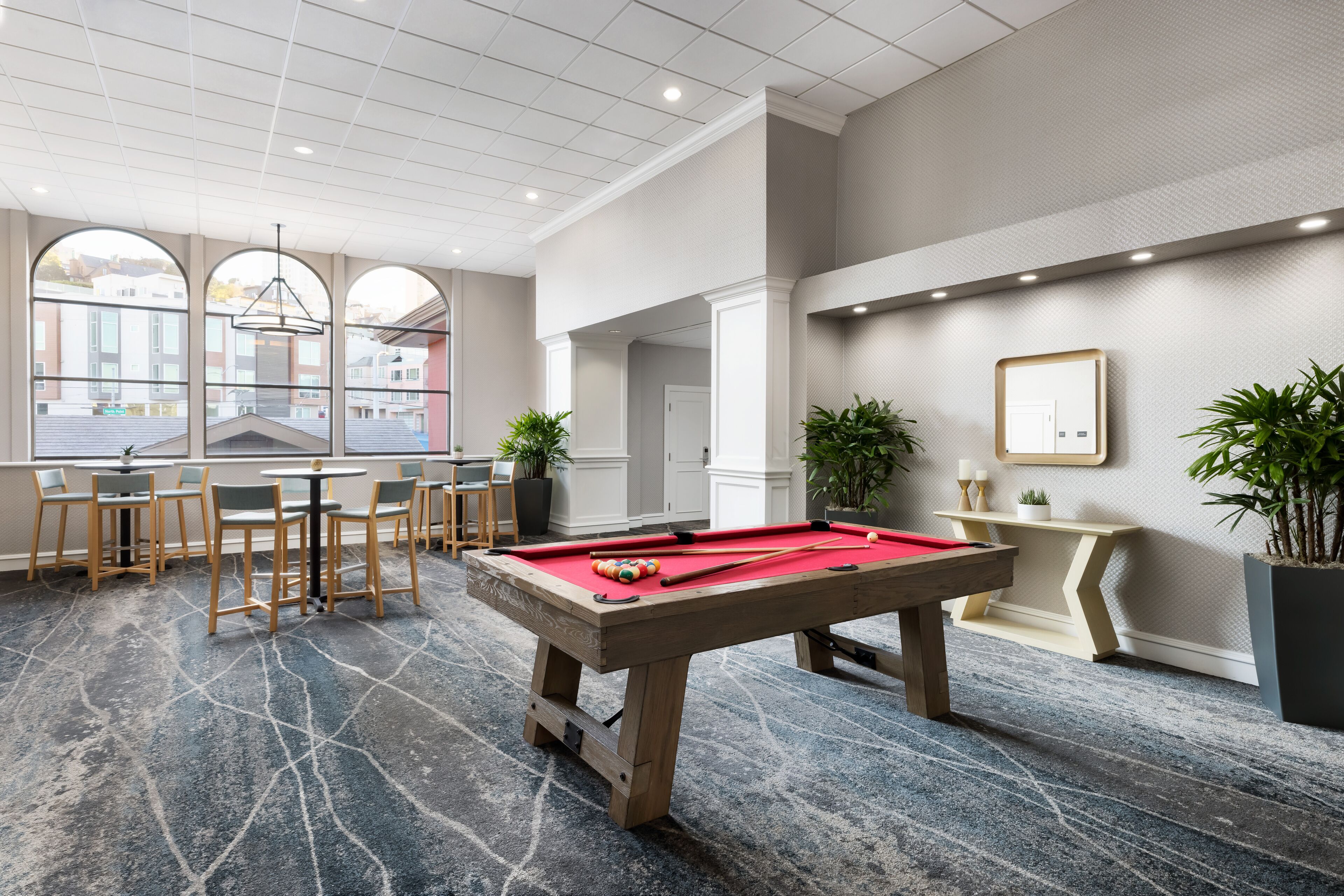 A billiards table in a modern room with a marble floor, seating area with chairs, and large windows offering views of buildings outside.