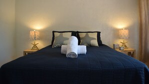 Een net opgemaakt bed met een donkerblauwe quilt, twee kussens in de vorm van zeesterren en een opgerolde witte handdoek in het midden. Er staan twee nachtkastjes met lampen en zeesterren decoraties.