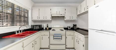 Una cocina compacta con armarios blancos, un lavamanos rojo, una estufa y un refrigerador.