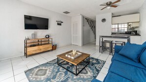 Een moderne woonkamer met een televisie, houten salontafel, blauwe bank en een keuken op de achtergrond.