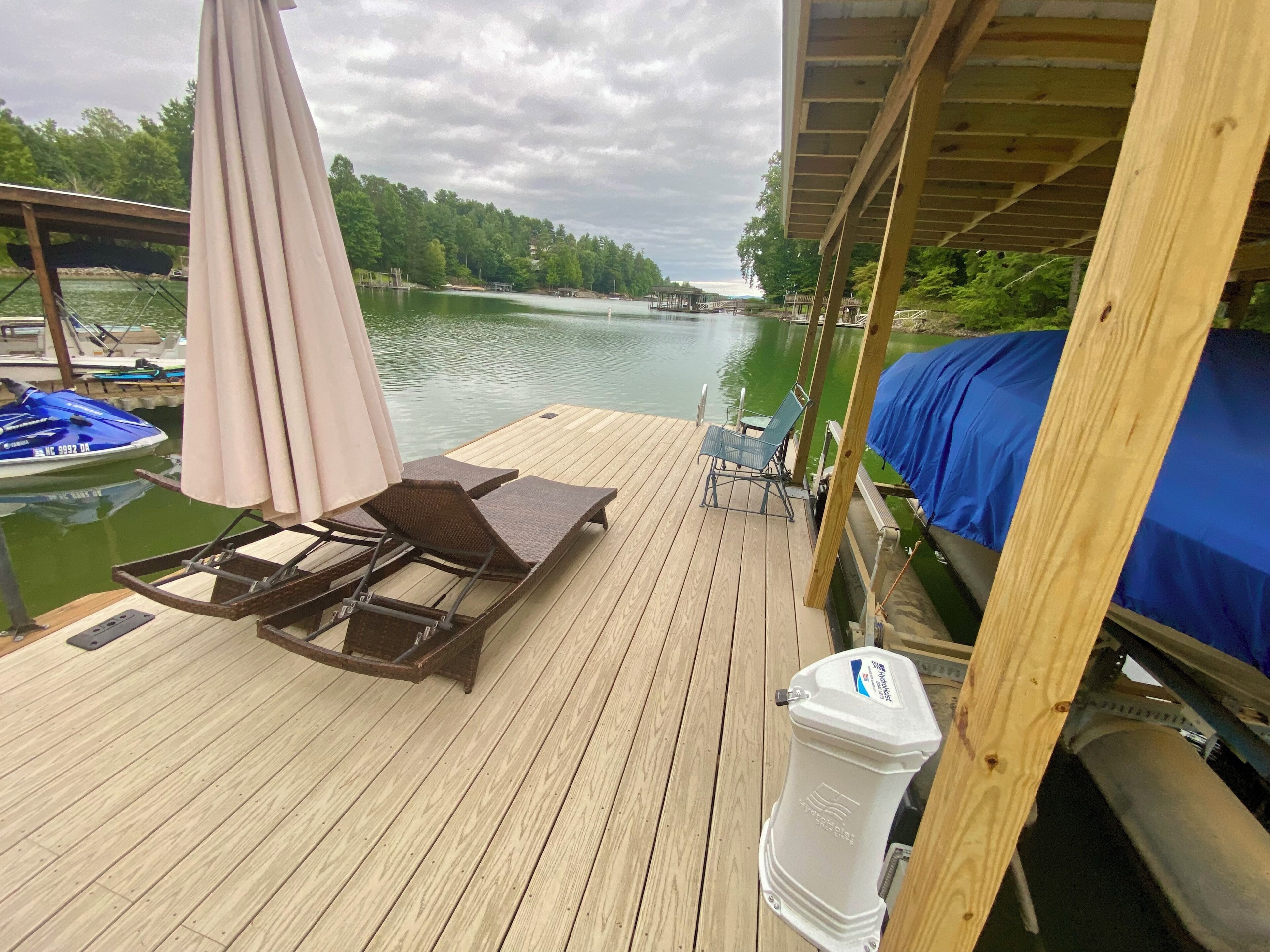 Een houten terras met lounge fauteuils en een tafel, met uitzicht op een rustige rivier met boten en een steiger op de achtergrond.