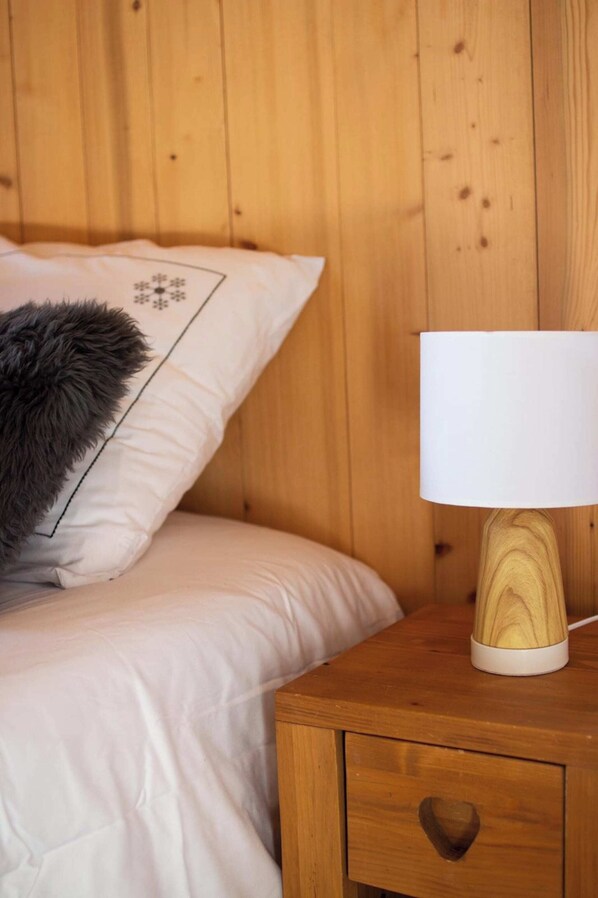 A wooden bed with a white pillow featuring a snowflake design and a fluffy gray pillow. A white lamp with a wooden base is on a wooden nightstand with a drawer. - COEUR DES ALPES - Edelweiss (Taninges)