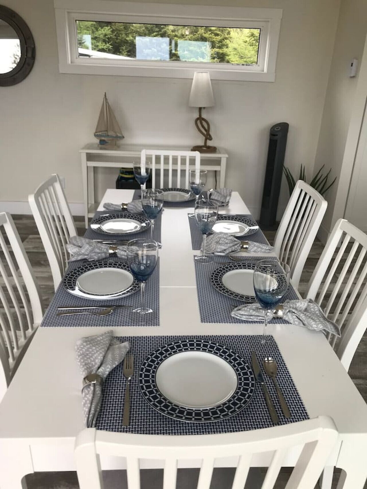 Ein Esstisch ist mit blauen Platzsets, weißen Tellern und Besteck gedeckt, umgeben von weißen Stühlen. Darüber befindet sich ein Fenster und eine kleine Tisch mit Lampe und dekorativen Gegenständen.