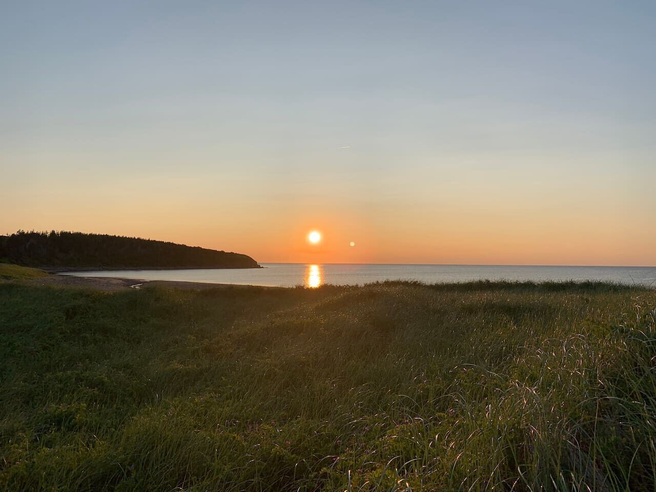 A sunset over a grassy field with a body of water and a distant landmass.