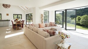 A spacious living room with a large sofa, a dining area with a table and chairs, and large windows offering a view of a forested area. - Set in the tranquil Teign Valley, Teign Vale makes the most of its position (Drewsteitgnton)