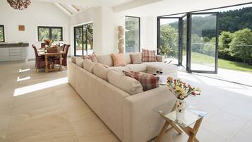 A spacious living room with a large sofa, a dining area with a table and chairs, and large windows offering a view of a forested area.