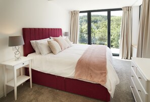A bedroom with a large bed, a nightstand, a dresser, and a view of a forest through a large window.