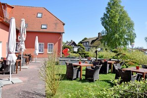 An outdoor cafe with tables and chairs, a red building in the background, and a tree. - 05 Double Room A (H) - The small hotel and apartments on Mönchgut! (Mönchgut)