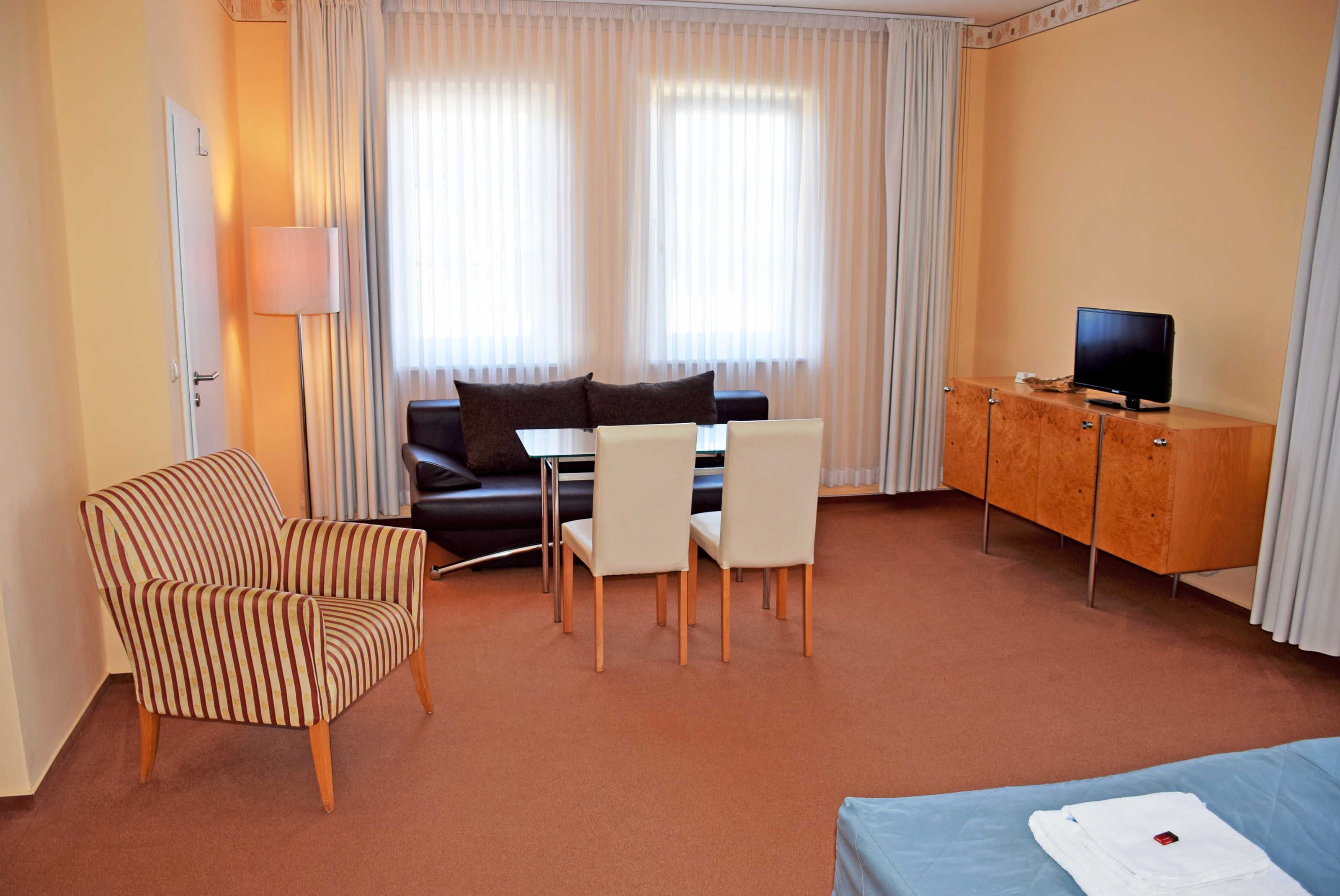 Une chambre d’hôtel avec un fauteuil à rayures, un canapé, une table à manger avec des chaises, un téléviseur posé sur un meuble en bois et un lit recouvert d’une couverture bleue.