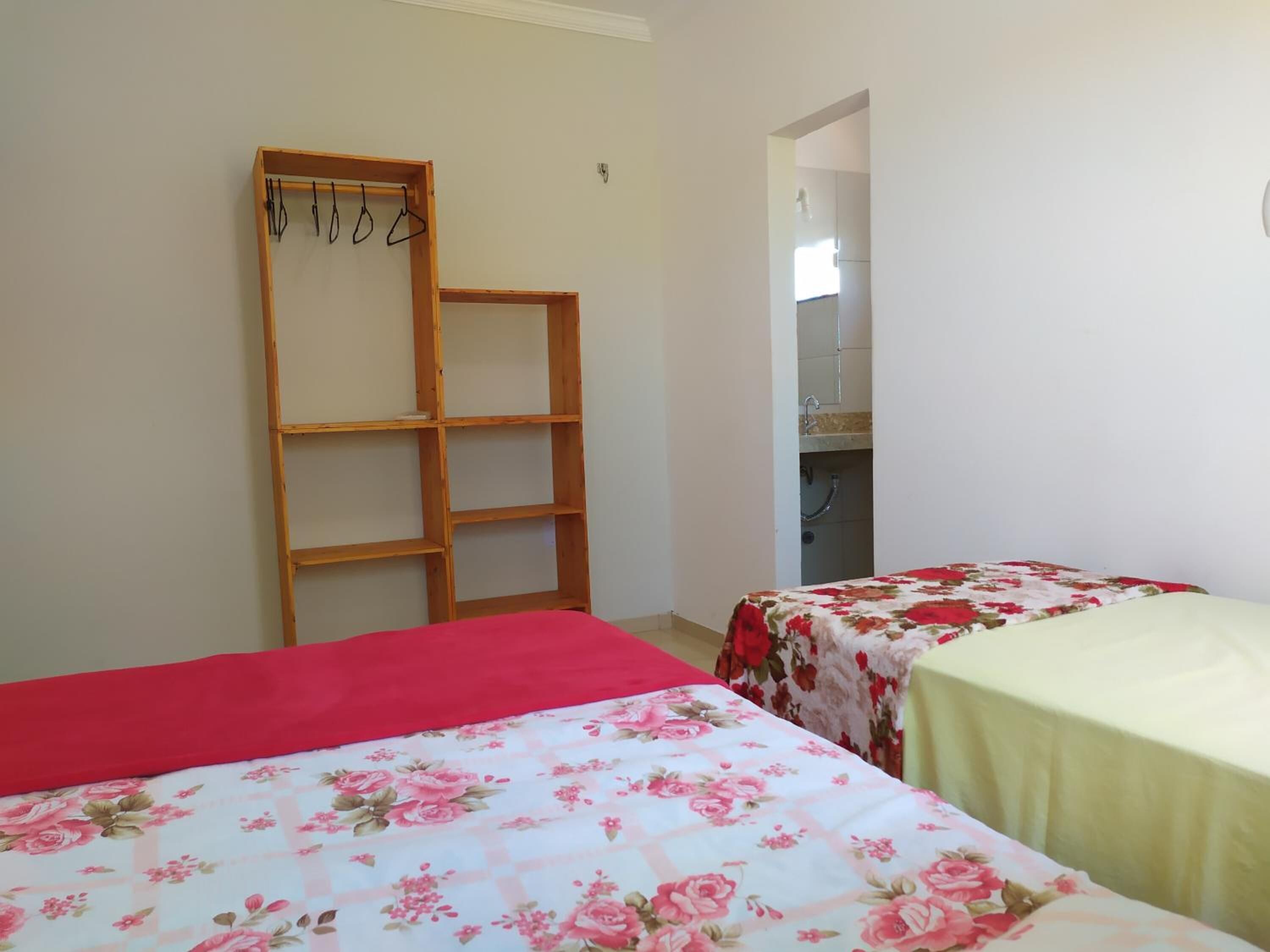 Quarto com duas camas, uma com colcha vermelha e outra com colcha floral. Há um armário de madeira com roupas penduradas e uma porta que dá para outro quarto.