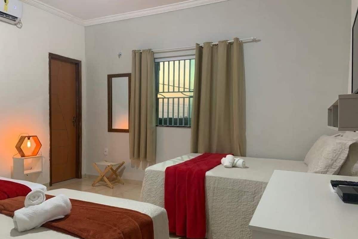Ein Hotelzimmer mit zwei Betten, einem kleinen Tisch, einem Stuhl, einem Fenster mit Gitterstäben und einer Lampe.
