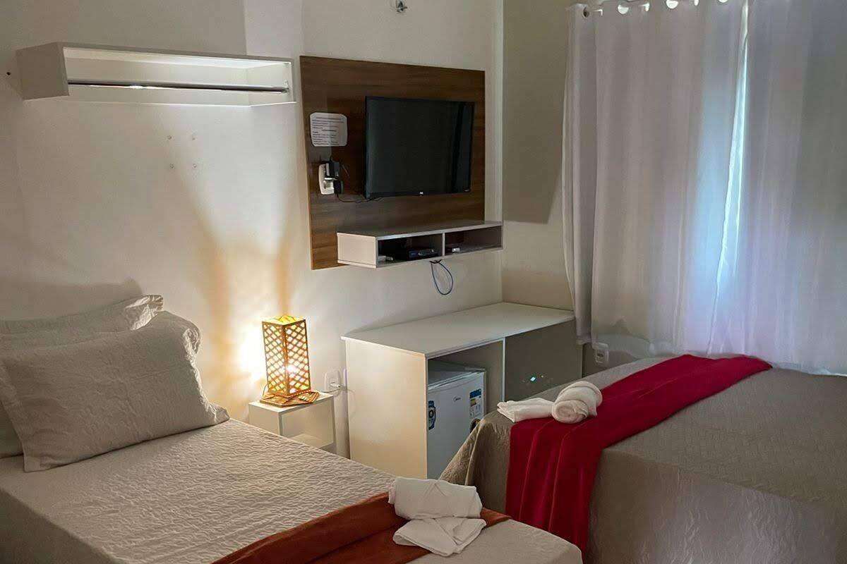 Ein Hotelzimmer mit zwei Betten, einem an der Wand montierten Fernseher, einem Schreibtisch mit Computer und einem Fenster mit Vorhängen.