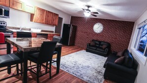 Una cocina con armarios de madera, un microondas y un refrigerador. Hay una mesa de comedor con sillas y una alfombra. Un reloj está en la pared.
