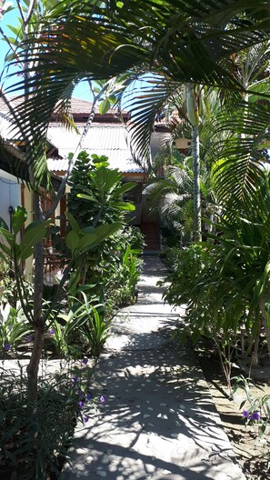 A narrow stone pathway flanked by lush tropical plants and a traditional building with a tiled roof in the background. - Sagita Bungalow (Gili Trawangan)