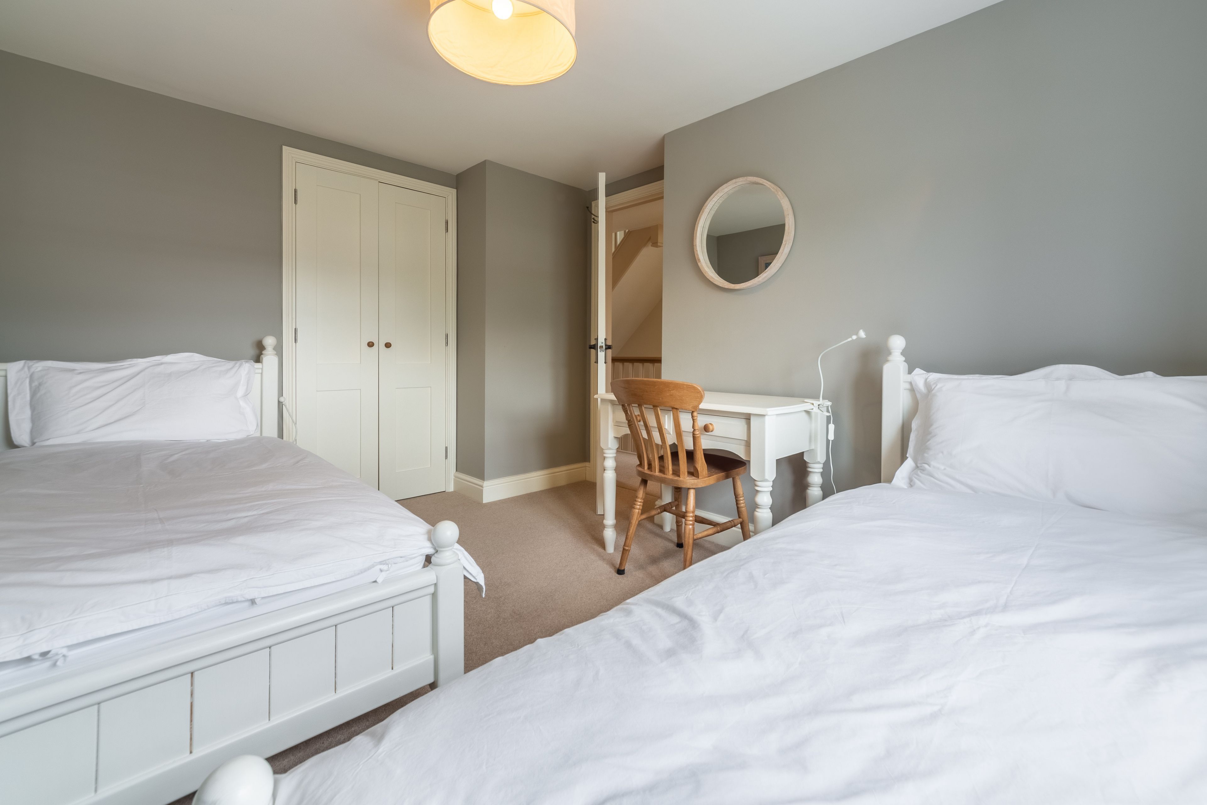 Une chambre avec deux lits simples, un bureau avec une chaise, un miroir rond et une porte donnant sur un escalier.