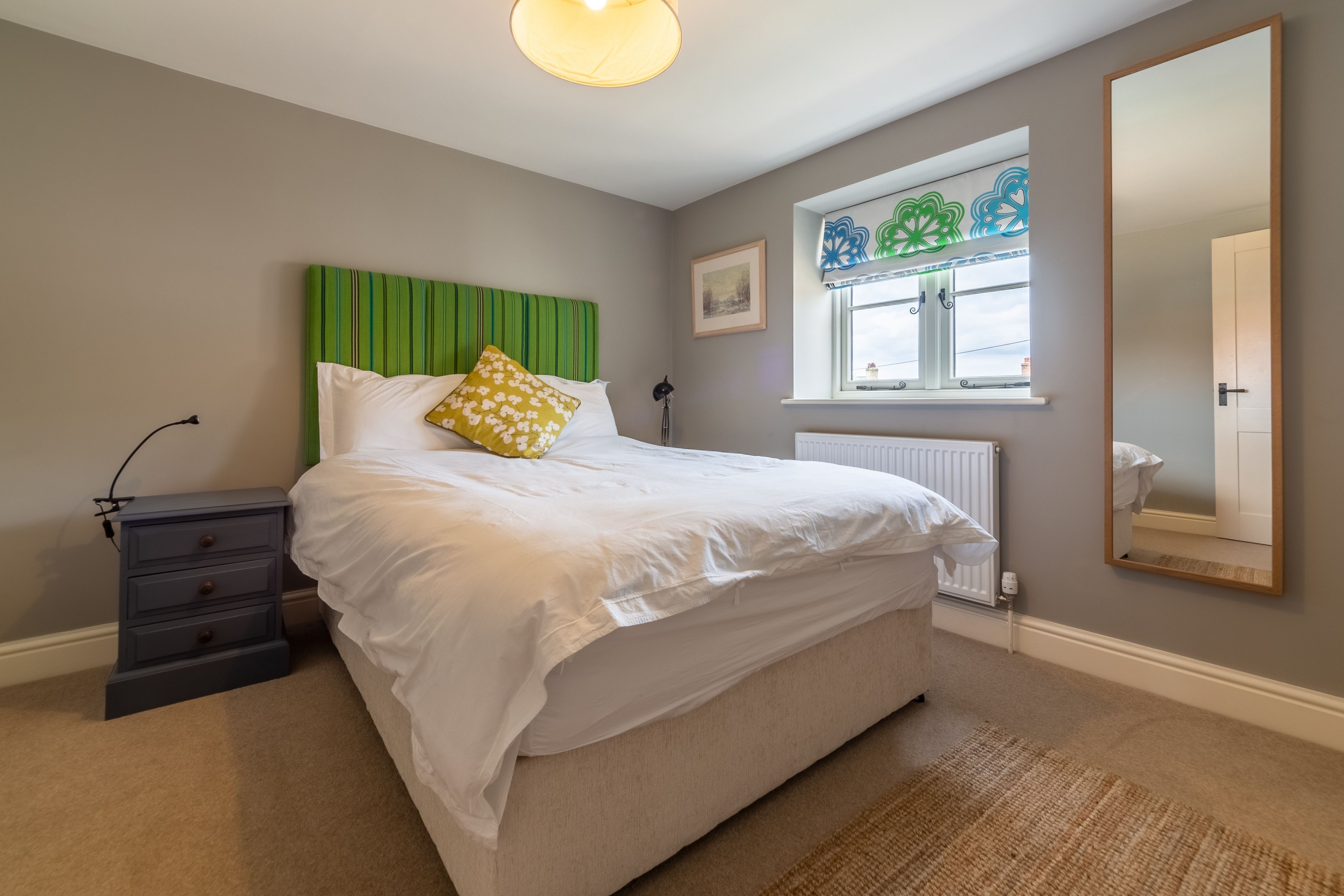 A bedroom with a bed, a nightstand, a window with decorative curtains, and a mirror on the wall.