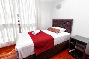 Cama bem arrumada com manta vermelha e travesseiro, mesa de cabeceira de madeira com luminária e janela com cortinas leves.