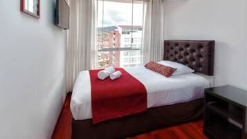 Habitación de hotel con una cama grande, una manta roja y vistas a edificios a través de la ventana.