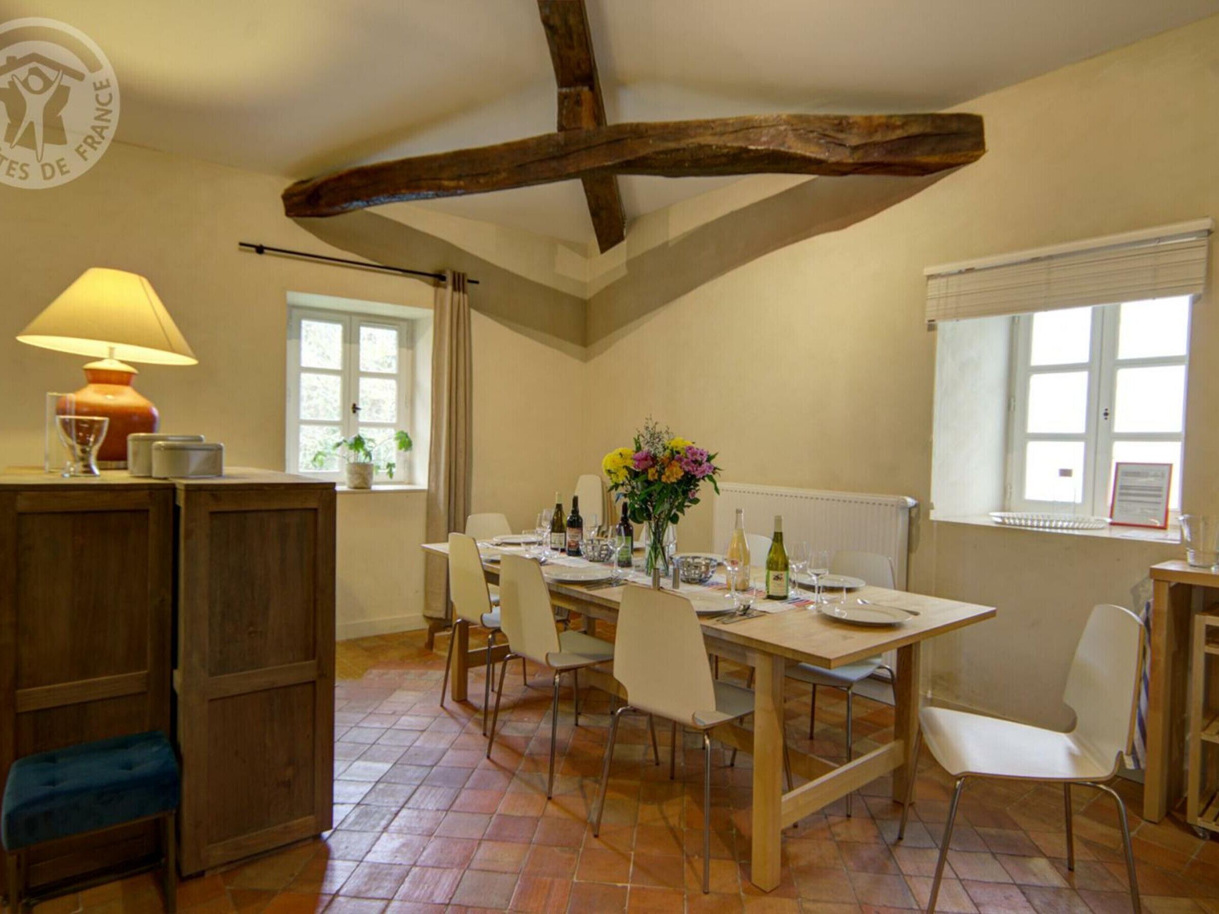 A dining area with a wooden table set for four, surrounded by white chairs. There is a lamp on a wooden cabinet, a window with curtains, and a radiator on the wall.