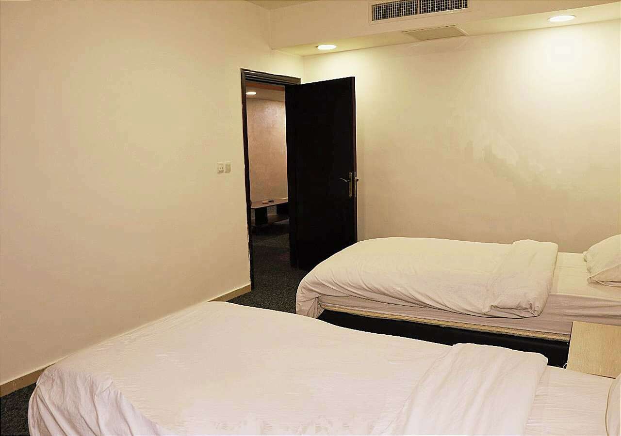 A small hotel room with two single beds, a door leading to a hallway, and a ceiling vent.