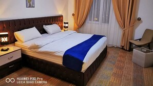 A hotel room with a large bed, a nightstand with a lamp, a chair, and a small table. - Skylight Apartments (Amman)