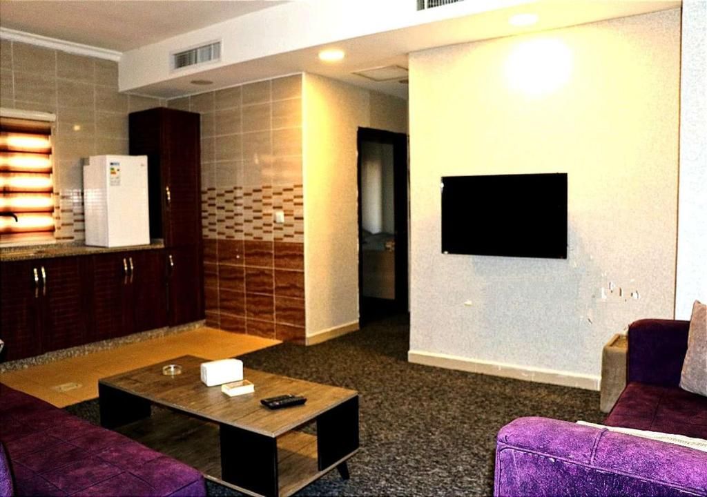 A modern living room with a sofa, a coffee table, a television mounted on the wall, and a kitchen area with cabinets and a refrigerator.