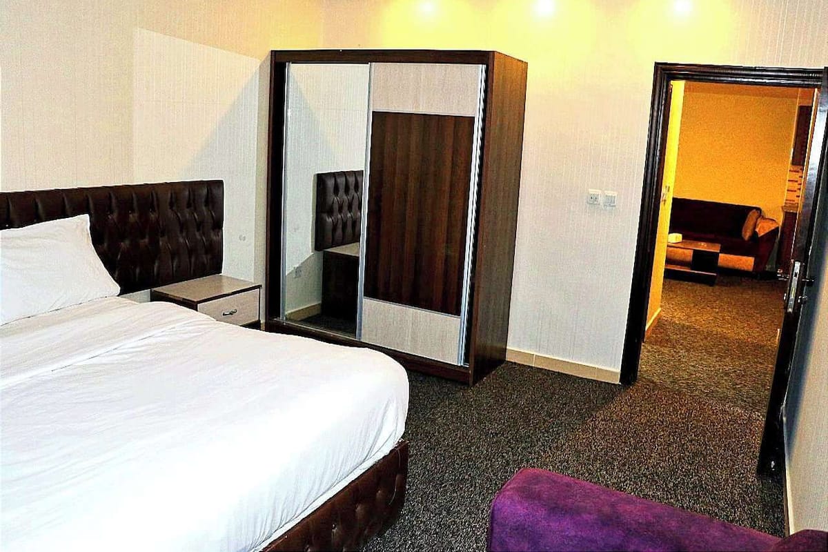 Una habitación de hotel con cama, armario, espejo y un reposapiés morado.