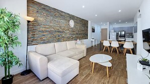 Ein modernes Wohnzimmer mit einer Eckcouch, einem Essbereich mit Tisch und Stühlen, einer Küche im Hintergrund und einer Wand mit einer Dekoration, inklusive einer Uhr.