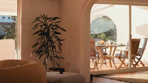 Ein Wohnzimmer mit einer Couch, einer großen Topfpflanze, einem Essbereich mit Stühlen und einem Tisch sowie einem Blick durch eine geöffnete Tür.
