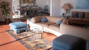 Ein Wohnzimmer mit Sofa, Hocker und einem Glastisch. Im Hintergrund ist eine Essecke mit Stühlen und einem Tisch zu sehen.
