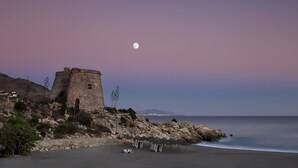 A coastal scene with a stone tower, beach umbrellas, and a calm sea under a moonlit sky. - Cotobro Rental House (Almuñécar)