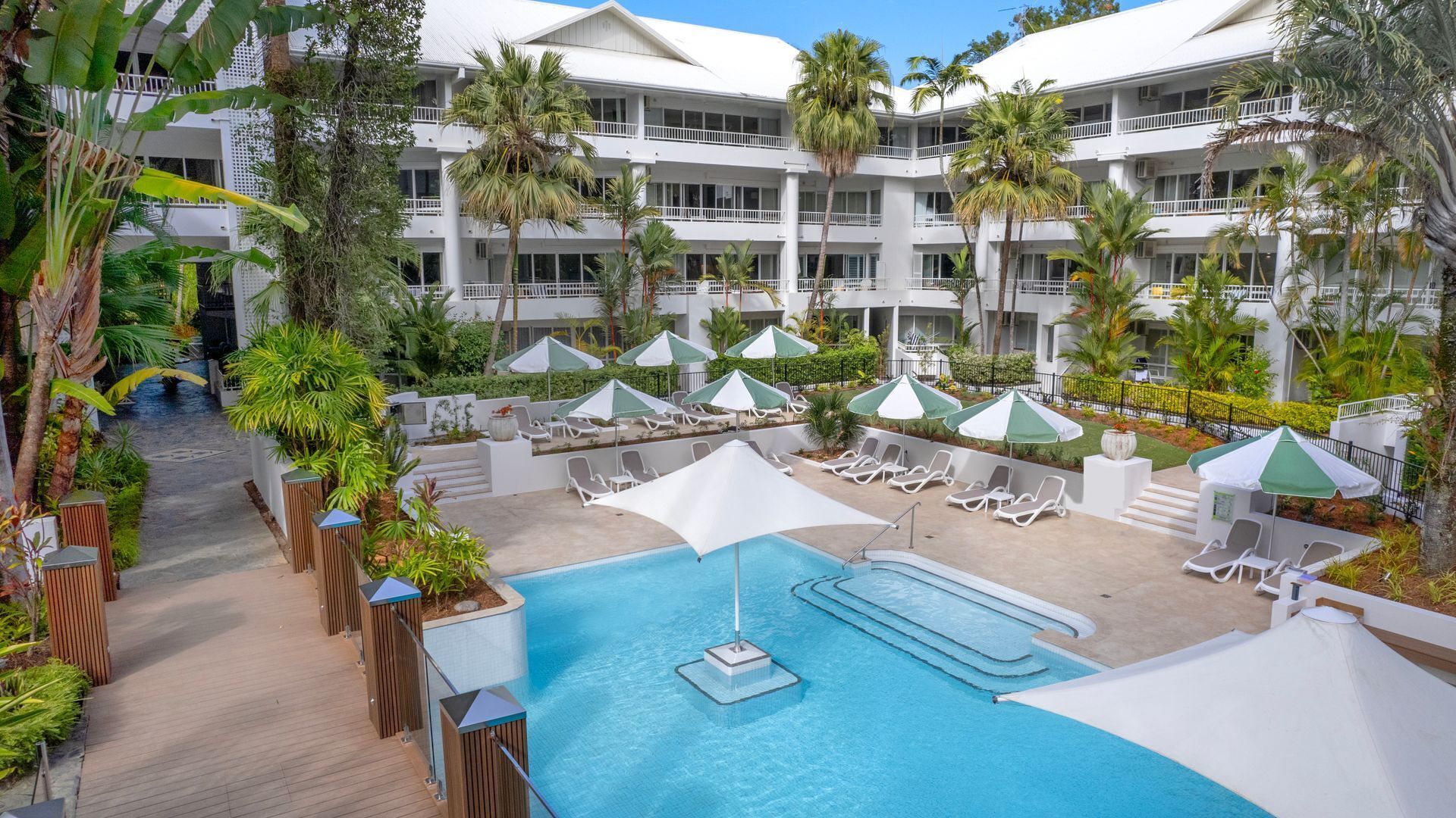 A hotel pool area with multiple lounge chairs and umbrellas, surrounded by lush greenery and a multi-story building.