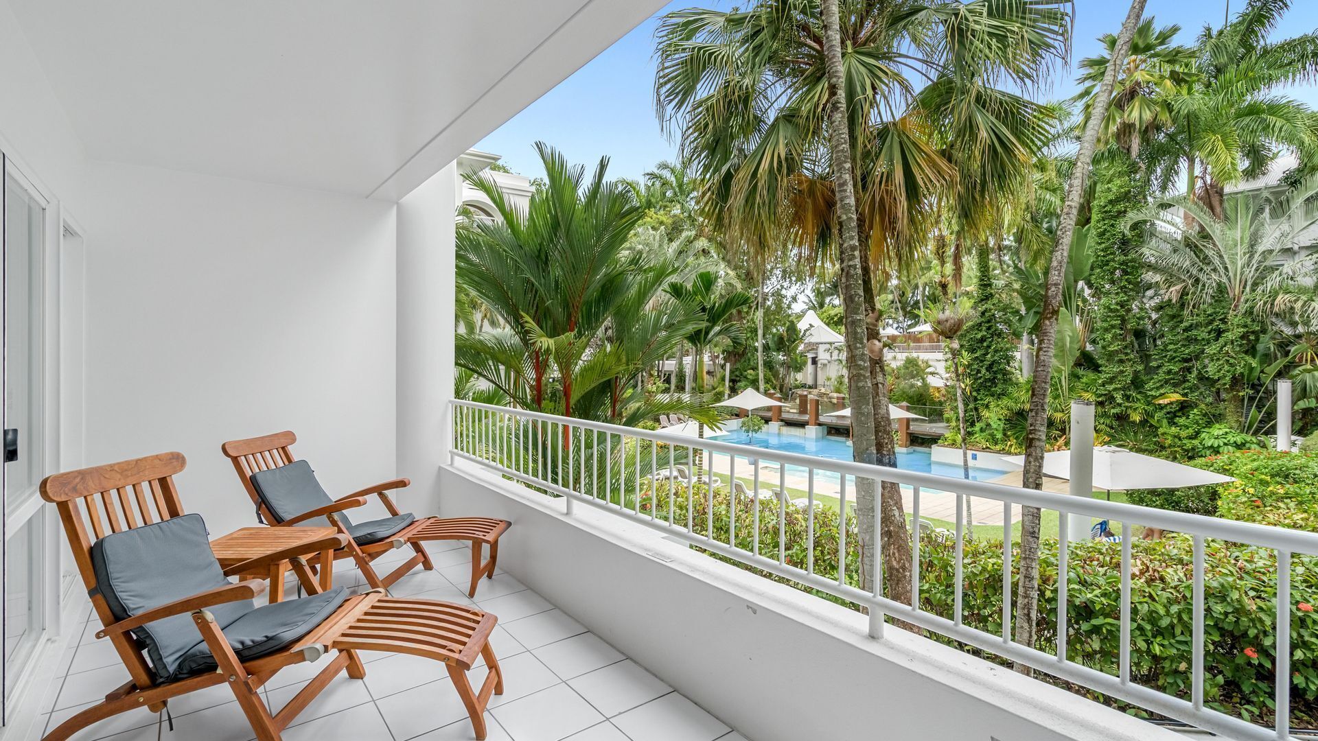 A balcony with wooden chairs and a view of a pool and tropical vegetation.
