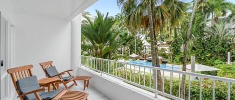 A balcony with wooden chairs and a view of a pool and tropical vegetation.