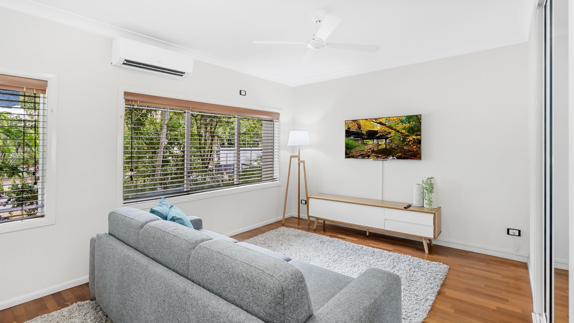 A modern living room with a grey sofa, a standing lamp, a TV mounted on the wall, and a window with blinds.