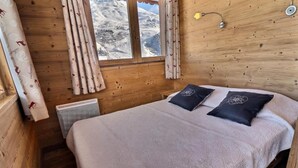 Una habitación de madera con una cama, cortinas, un radiador y una ventana que ofrece vistas nevadas.