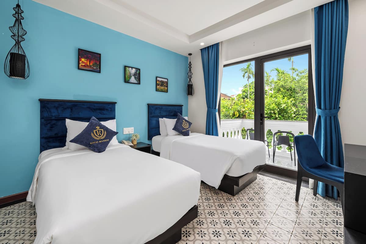 A hotel room with two beds, a balcony with a view of greenery, and a patterned floor.