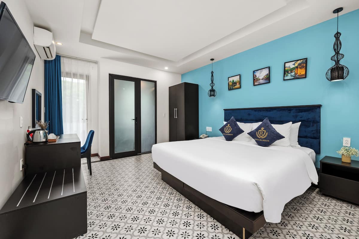 A modern hotel room with a large bed, a desk, and a chair. There is a wardrobe, a TV, and a window with curtains.