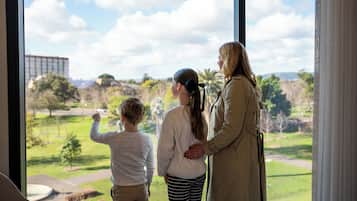 A family of three standing by a window overlooking a park with trees and a building in the distance.