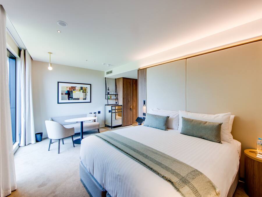 A modern hotel room with a large bed, a desk, and a chair. There is a small table with water bottles on it.