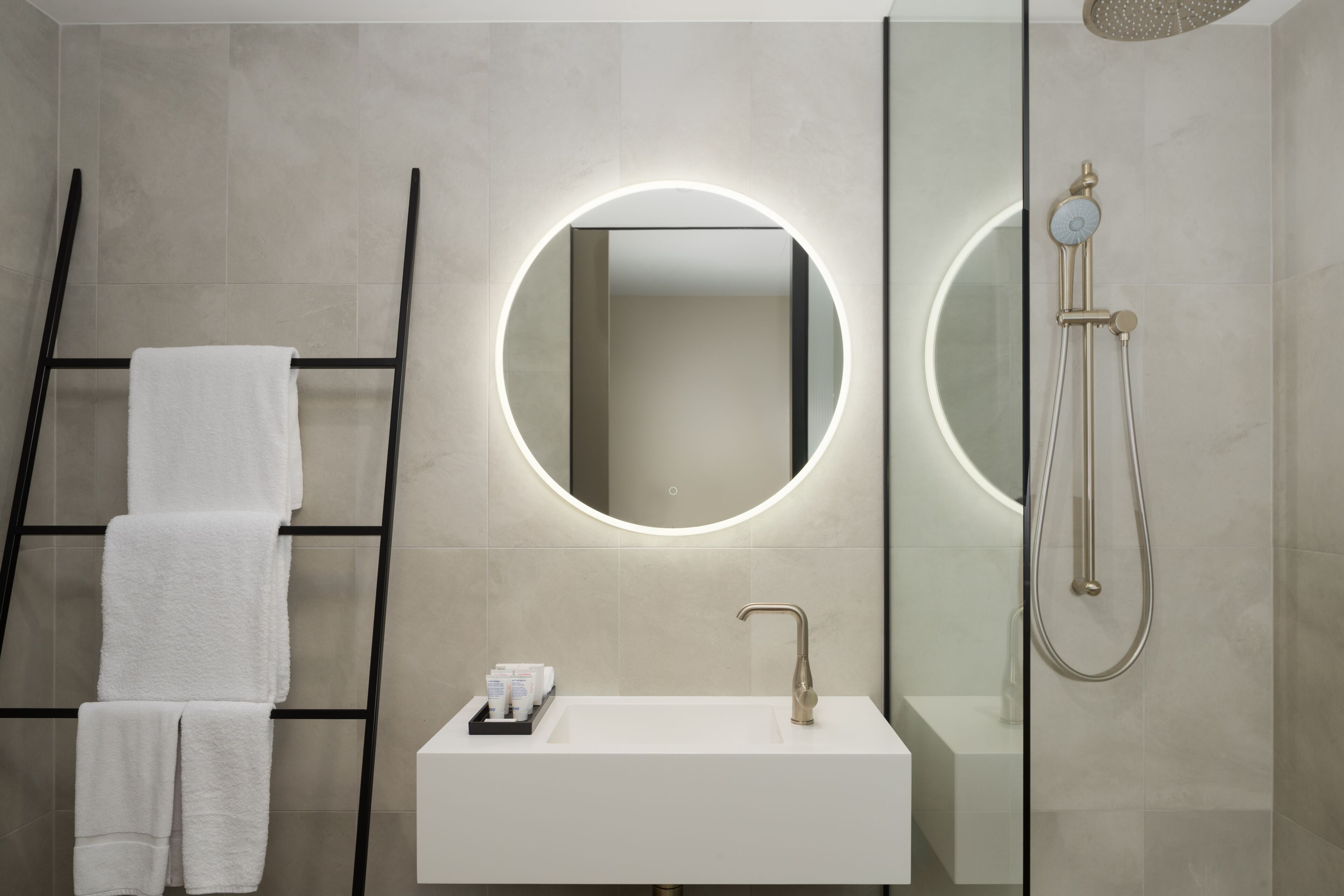 A modern bathroom with a white sink, a round mirror with a light, a shower, and a towel rack with towels.
