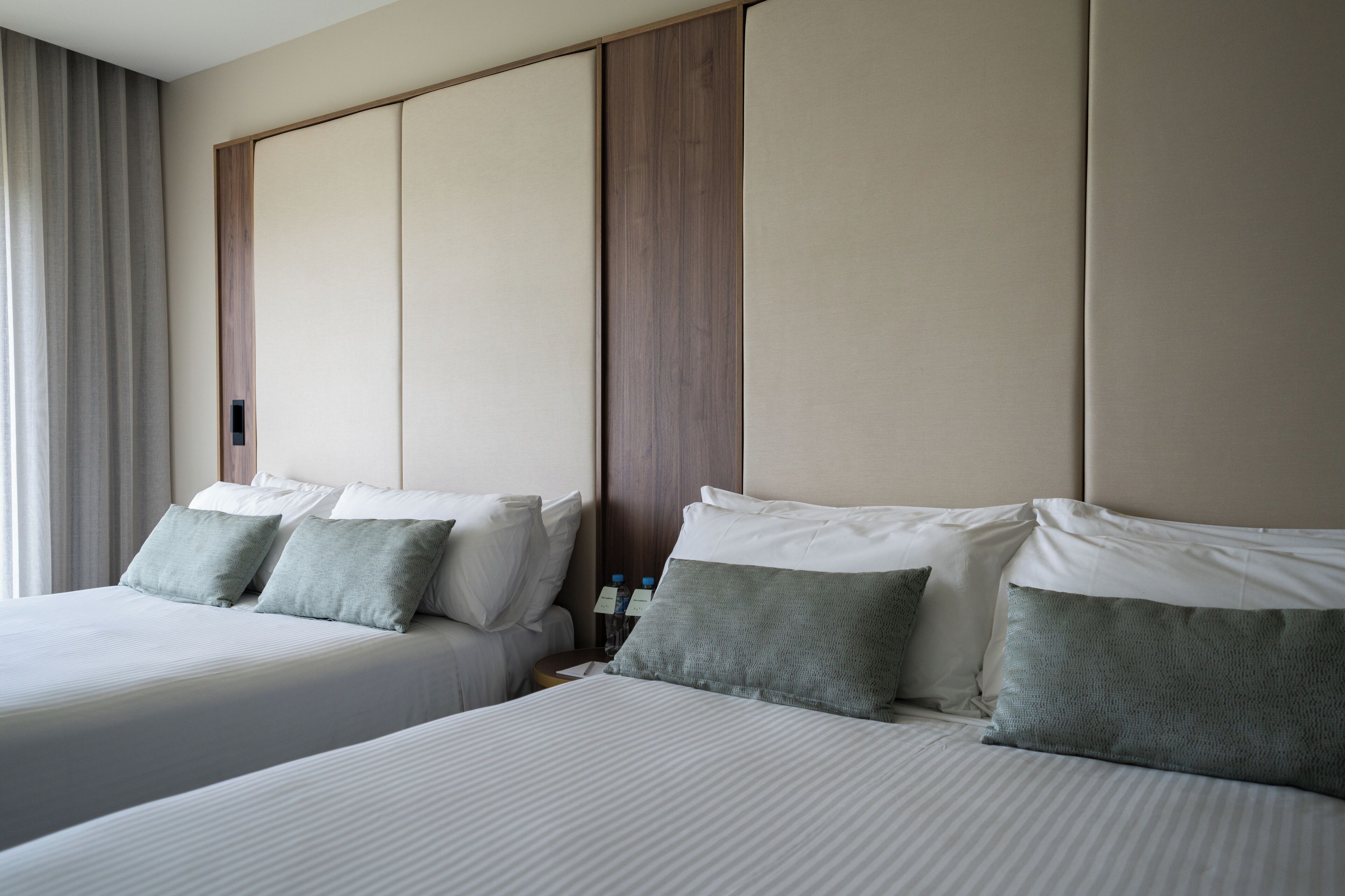 Ein Hotelzimmer mit zwei Betten, jeweils mit weißer Bettwäsche und grünen Kissen. Es gibt ein Holz-Kopfteil und einen Nachttisch mit Lampe und Wasserflaschen.